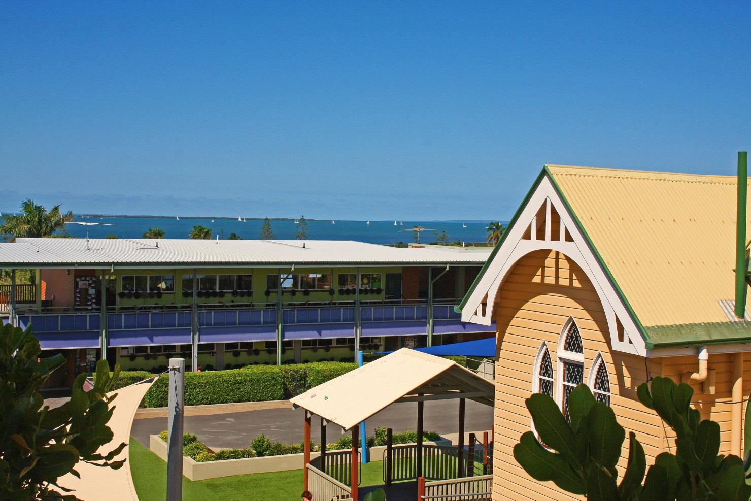 School courtyard with a playground, shaded walkways, and classroom buildings, with the ocean visible in the background.