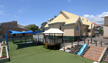School playground with climbing equipment, slides, and shade structures beside a heritage‑style building.