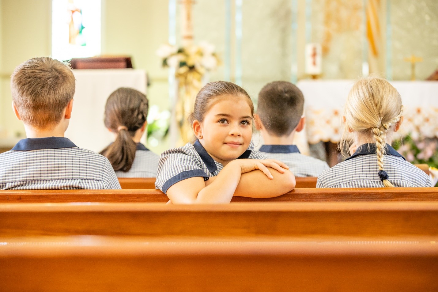 Student seated in a church pew, with other students in uniform facing the altar in the background.