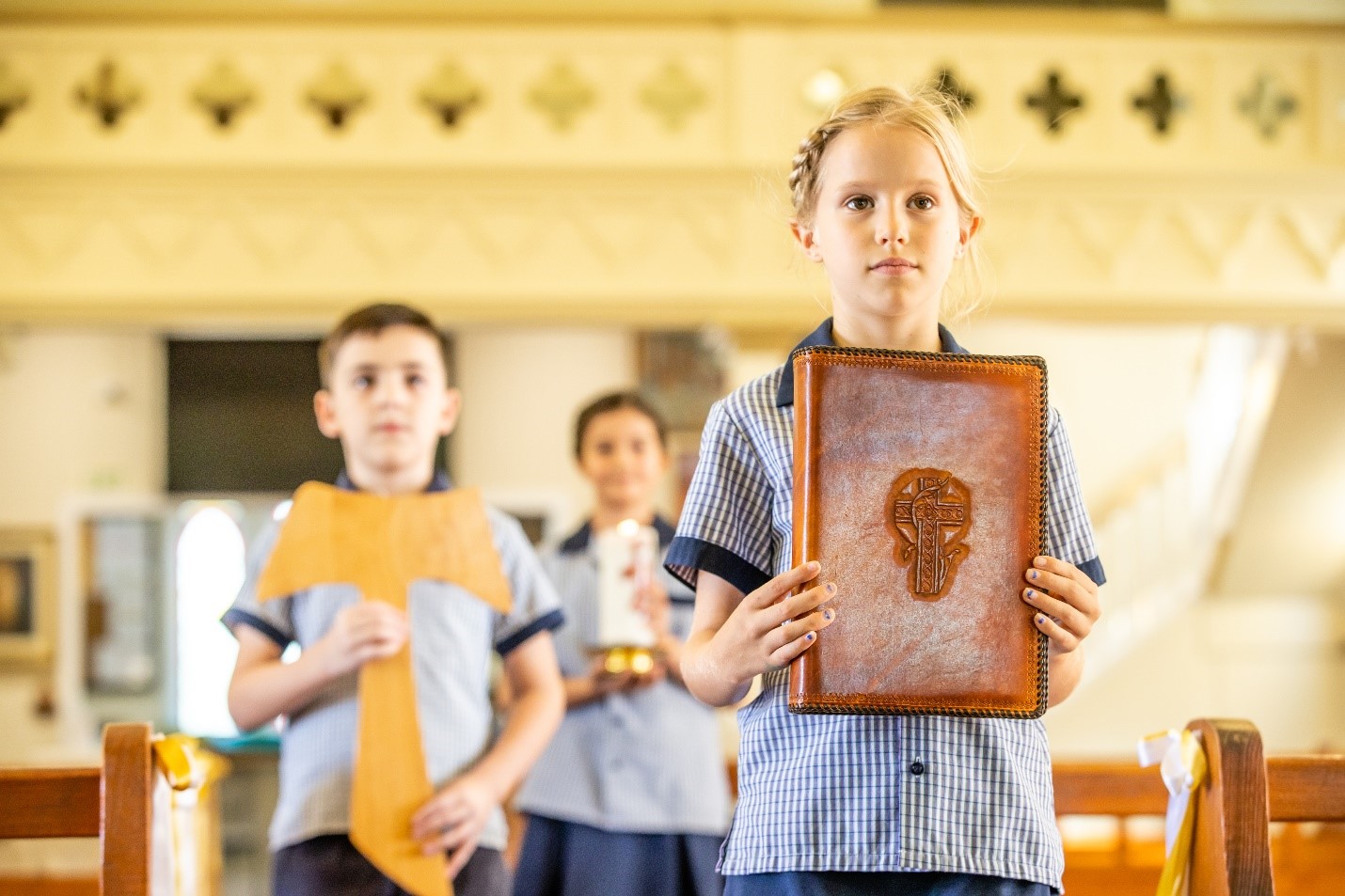 Students in school uniform standing in a church, with one holding a large book and others holding a candle and cloth.
