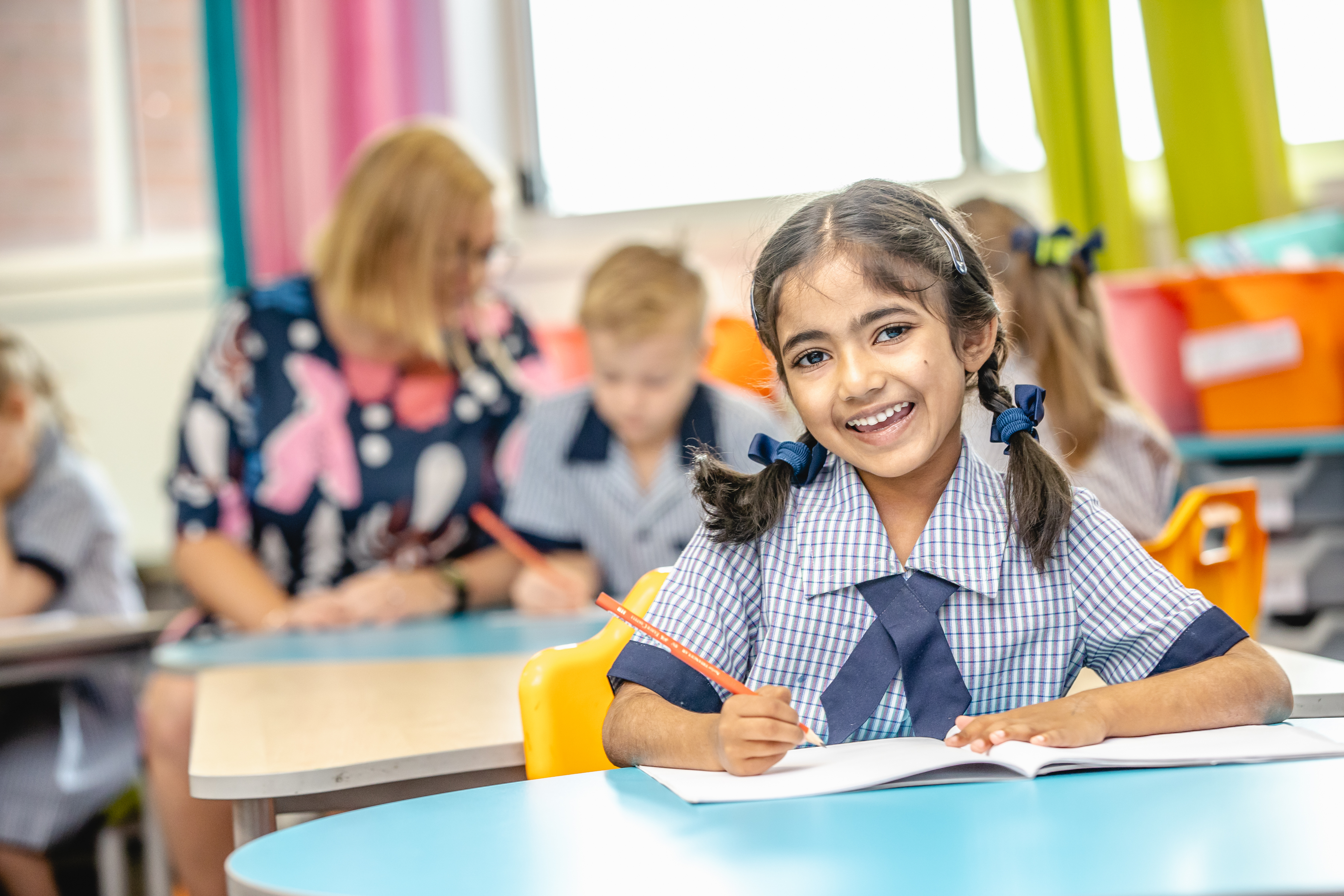 Student in a classroom writing in a notebook, with other students and a teacher in the background.