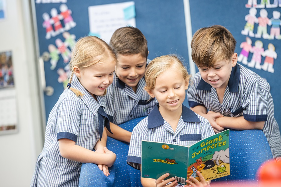 Students in uniform gathered around, reading a book together in a classroom.