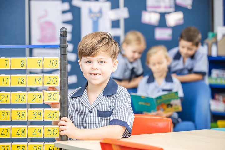 Student in a classroom using a number chart and counting blocks, with other students reading in the background.