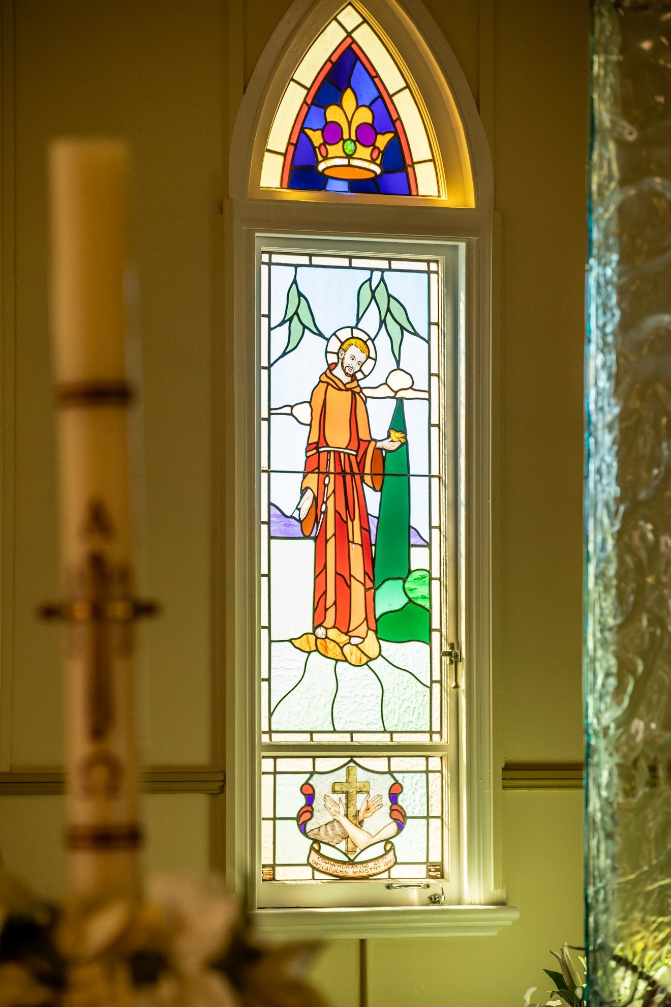 Stained glass window of a saint holding a bird, with a crown above and a commemorative panel below.